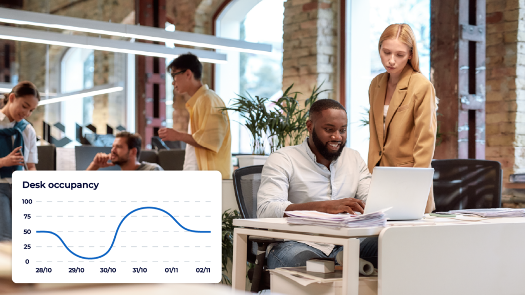 A busy office with two people looking at a laptop to gain insights on workplace analytics and desk occupancy